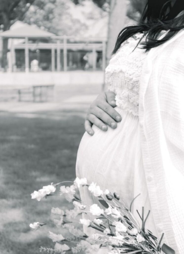 Maternity photo of client in white dress holding flowers outdoors.